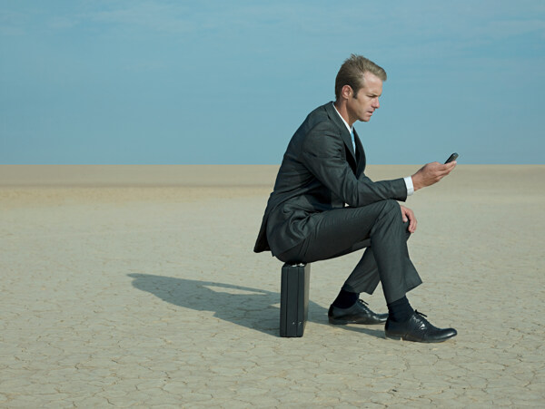 在沙漠中坐着皮箱玩手机的外国商业男人图片